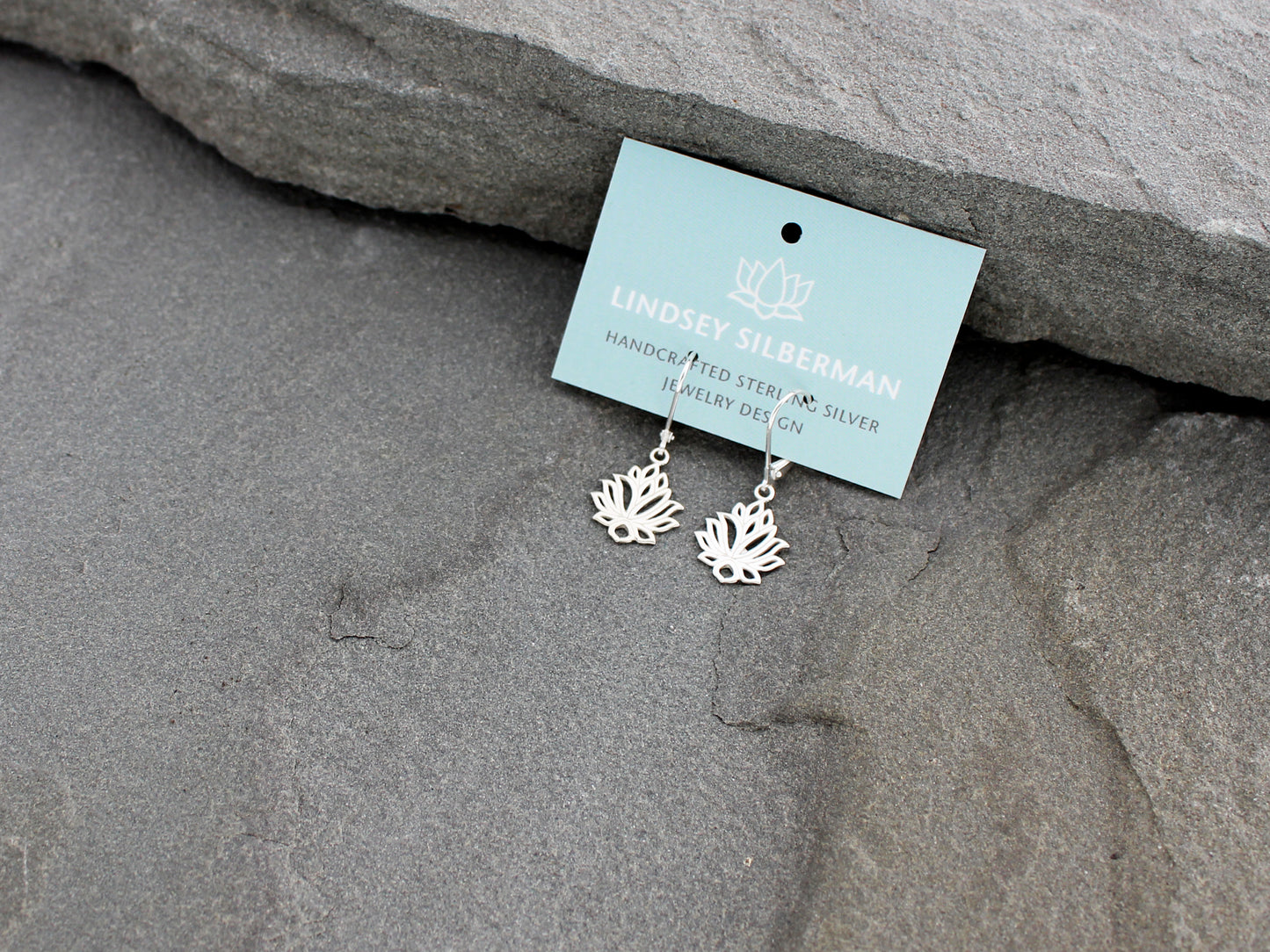 Silver lotus flower earrings on a card with 'Lindsey Silberman' branding, placed on gray stone.